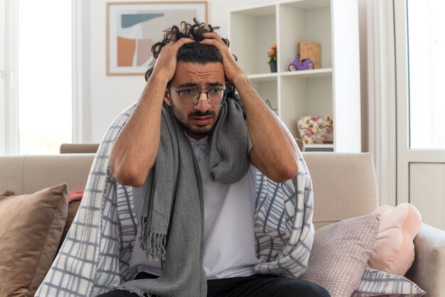 anxious young ill caucasian man in optical glasses wrapped in plaid with scarf around his neck putting hands on his head sitting on couch at living room